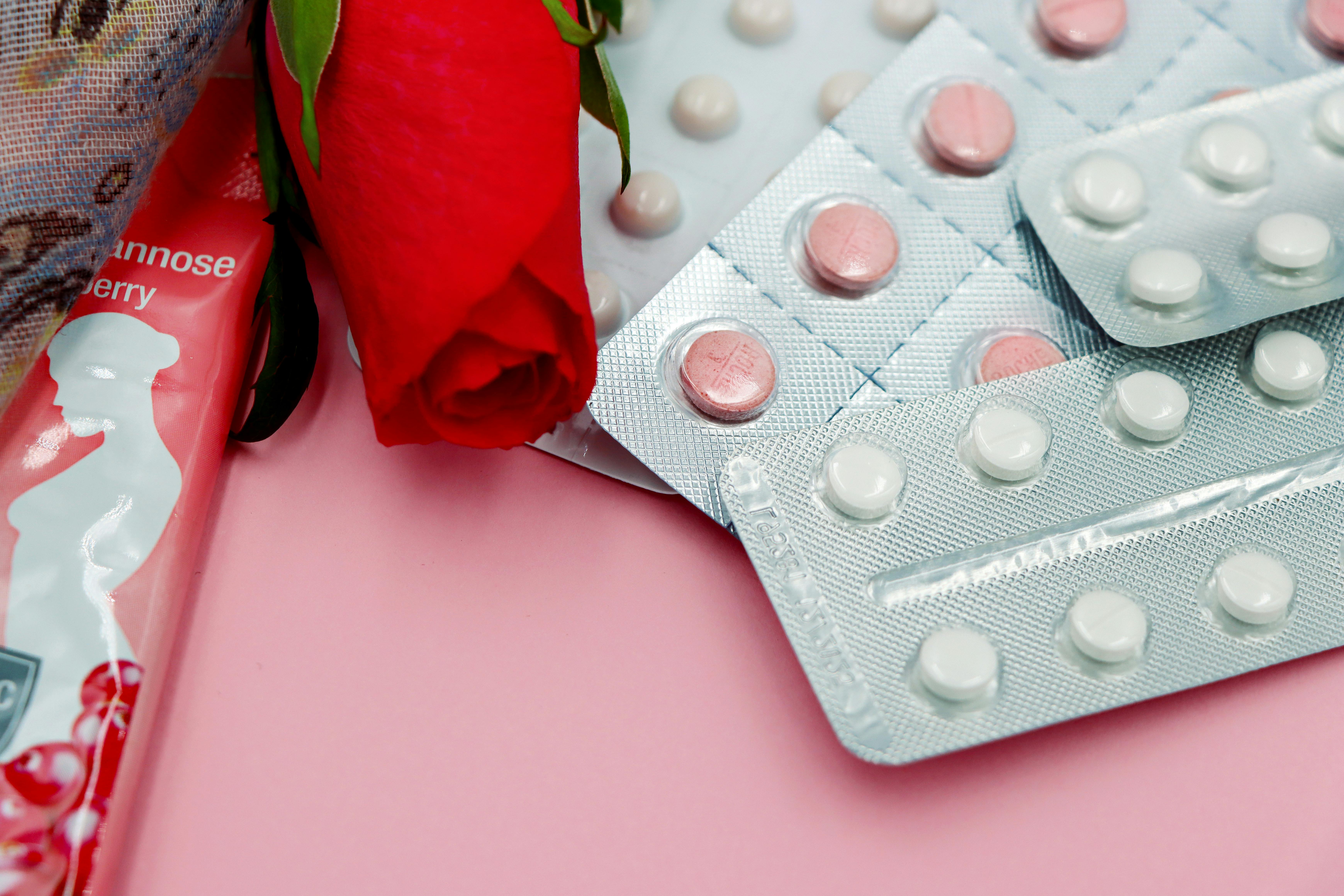 Blísteres de pastillas anticonceptivas junto a una rosa roja y un envoltorio femenino sobre fondo rosa, simbolizando el tratamiento tradicional del síndrome de ovario poliquístico (SOP)